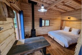 Intérieur chaleureux tout en bois d'un hébergement insolite aux Cabanes du Lac de Pierre-Percée en Meurthe-et-Moselle, révélant une chambre douillette avec un grand lit double, une table rustique et un poêle à bois.