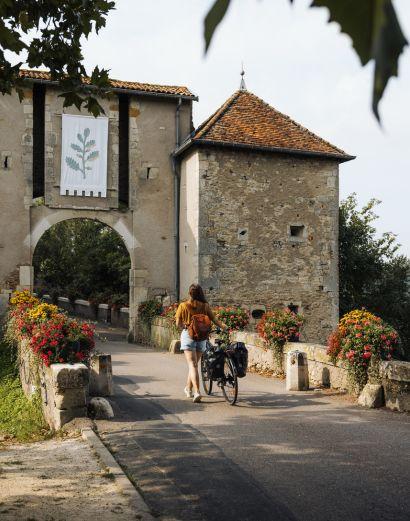 Une cycliste avec sacoches marche sur un pont fleuri à l'entrée de la cité médiévale de Liverdun en Meurthe-et-Moselle. Elle passe devant une ancienne porte fortifiée et une tour en pierre défensive, avec un drapeau orné d'un blason sur la façade.