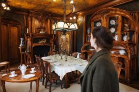 Une jeune femme regarde l'intérieur d'une pièce de la Ville Majorelle à Nancy, on y voit notamment du mobilier en bois.